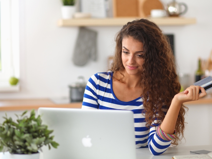 A beautiful woman with long brown, wavy hair sitting in front of her laptop wearing a blue and white striped shirt and holding out a credit card in her left hand.