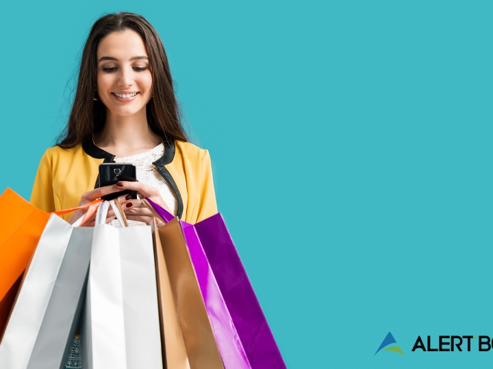 A beautiful woman with brow n hair holding and looking at her cellphone, with five shopping bags also in her hands. Text on the image reads "6 Tips to Prepare Your E-Commerce Site for the Biggest Holiday Traffic Surge Ever"