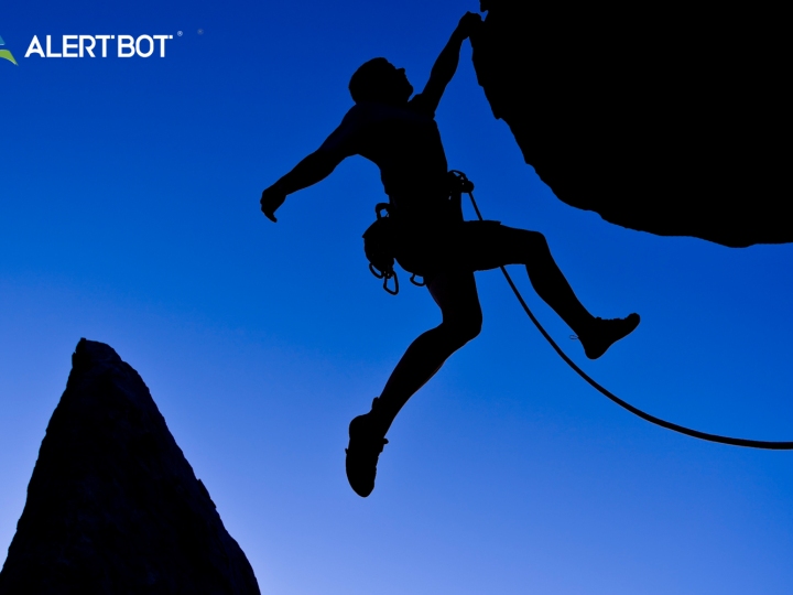 A mountain climber is silhouetted on a deep blue sky background as he hangs off a cliff by one hand, and a tether hangs off his belt.