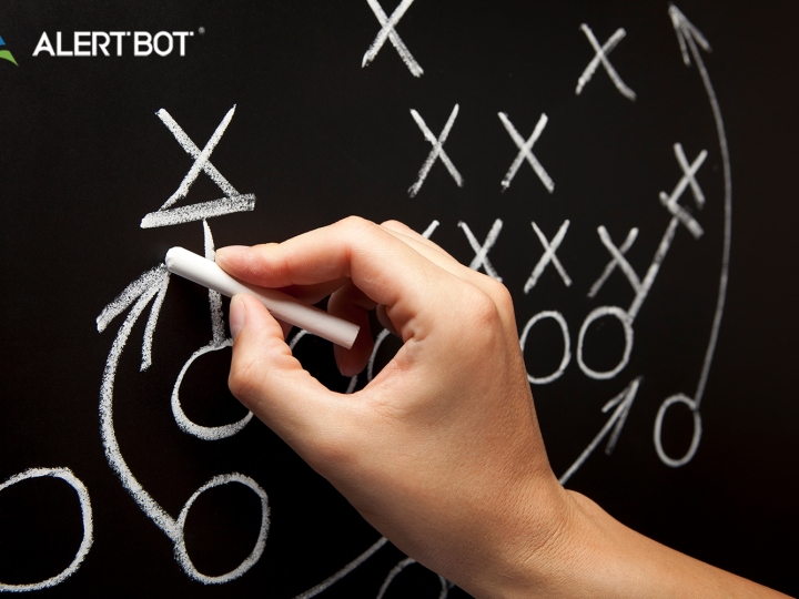 A hand holding chalk writes football plays on a blackboard, using x's, arrows and o's.